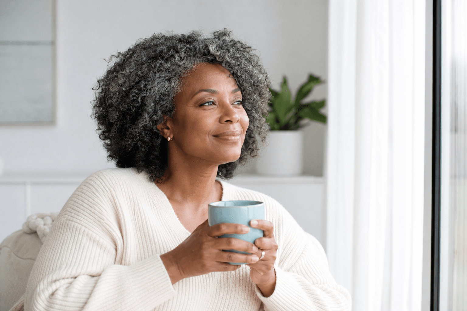 A confident Black woman around 50 holds an aqua mug by a bright window, looking rested and content